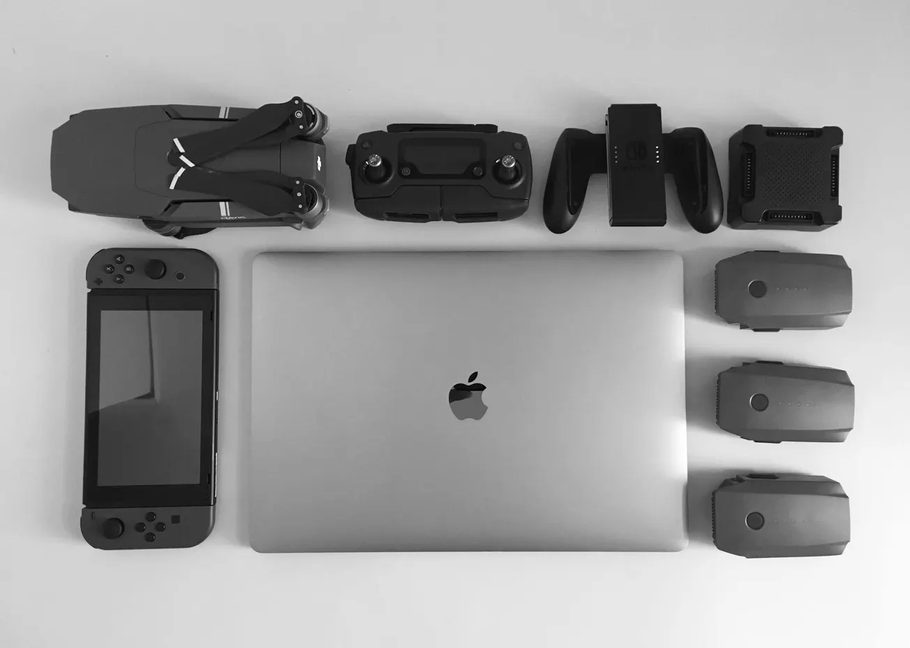 Various tech gadgets arranged neatly on a white surface. Includes a closed laptop with an Apple logo, a Nintendo Switch, a drone, controller, and four battery packs.