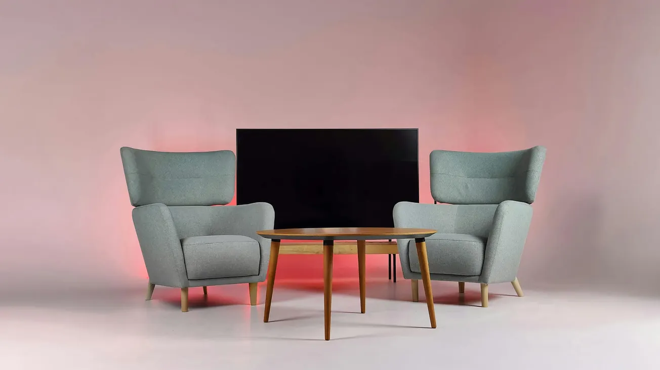 Two gray armchairs face each other with a round wooden coffee table between them. A flat-screen TV sits behind, against a soft pink background.
