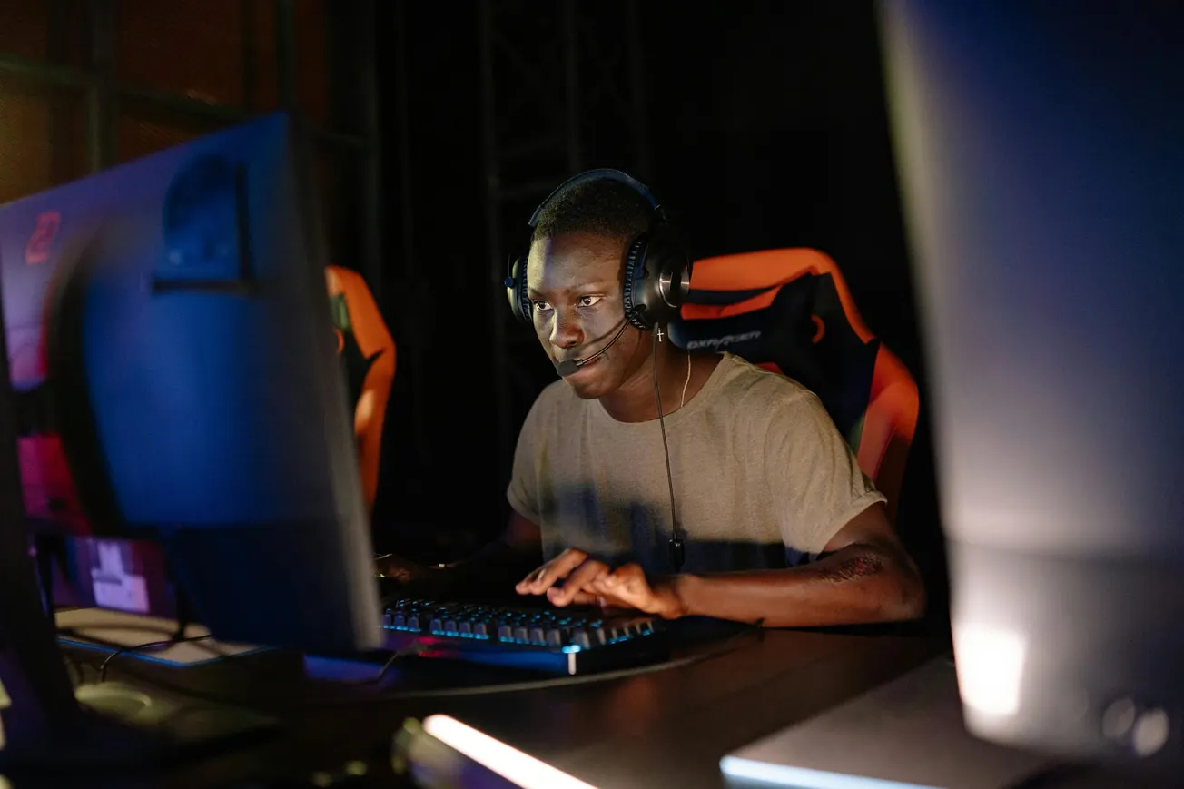 A person wearing headphones intensely focuses on a computer screen in a dimly lit room. They sit in a gaming chair, creating a concentrated gaming atmosphere.