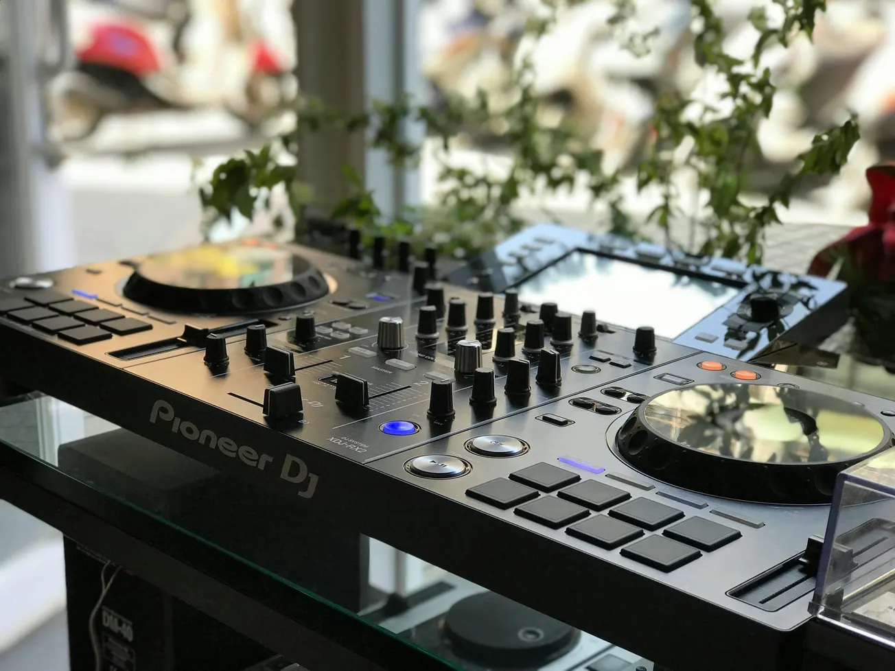 DJ controller on a reflective surface, featuring knobs, pads, and illuminated buttons. Background shows a blurred street scene and greenery.