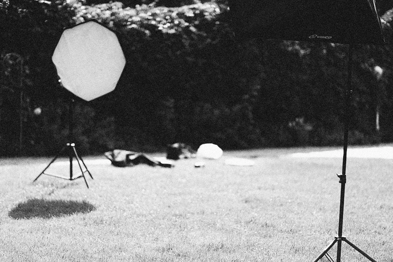 Black and white photo of a grassy outdoor setting with studio lighting equipment, including softboxes, under a canopy of trees, suggesting a photo shoot.