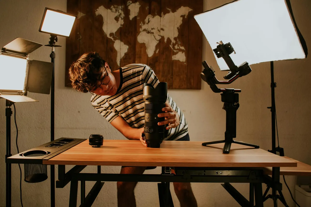 A person in a striped shirt examines a large camera lens on a wooden table, surrounded by professional lighting.