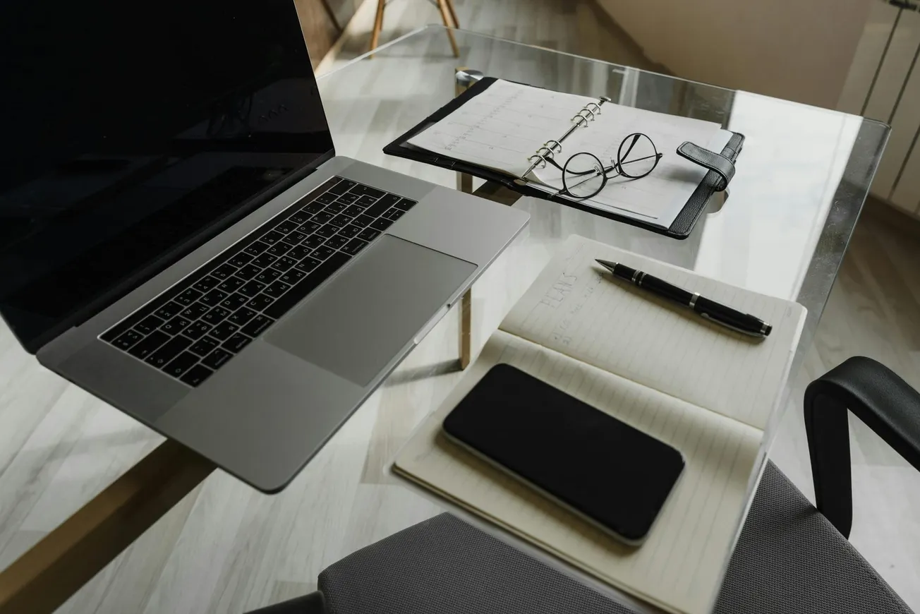 A glass-top desk features a laptop, an open notebook with a black smartphone, a pen, a planner, and eyeglasses. The scene conveys a minimalist, organized workspace.
