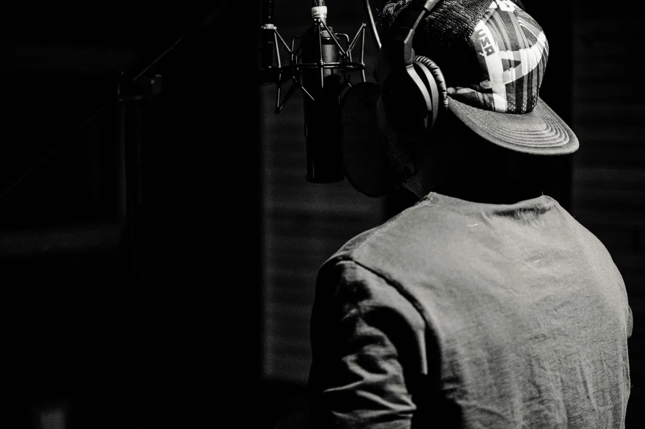 Black and white image of a person wearing a cap and headphones, facing a microphone in a recording studio. Dim lighting creates a focused, intimate mood.