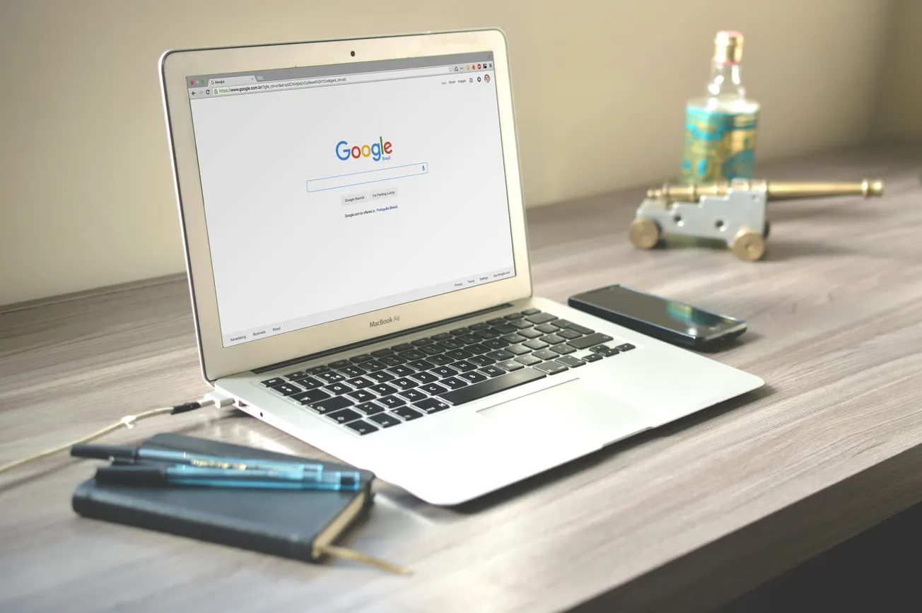 A laptop displaying the Google homepage sits on a wooden desk, alongside a notebook, pen, smartphone, and decorative bottle with a cannon model.