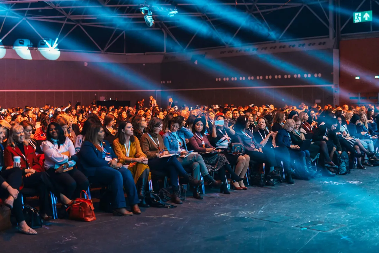 Audience at a conference in a large hall. People of diverse backgrounds sit attentively under blue spotlights, creating an energetic and engaged atmosphere.