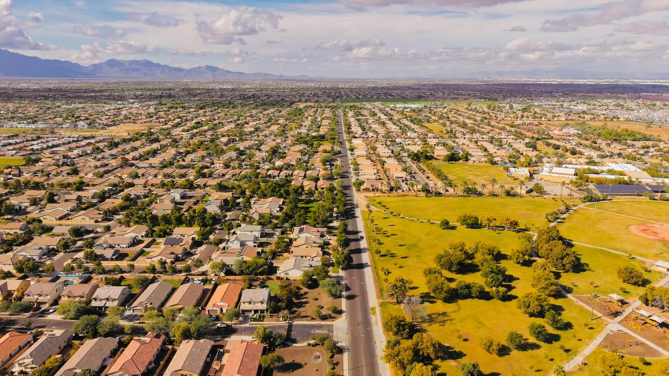 Aerial view of a sprawling suburban area with a grid layout. Houses with red-tiled roofs on the left, green open fields on the right, under a blue sky.