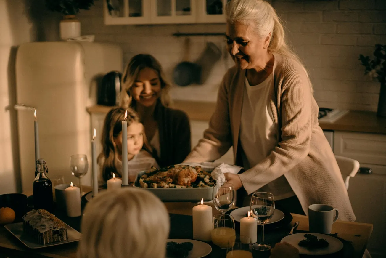A woman with white hair joyfully serves a roasted dish at a warmly lit dinner table. Family, including a child, sit eagerly, surrounded by candles. Cozy and festive atmosphere.
