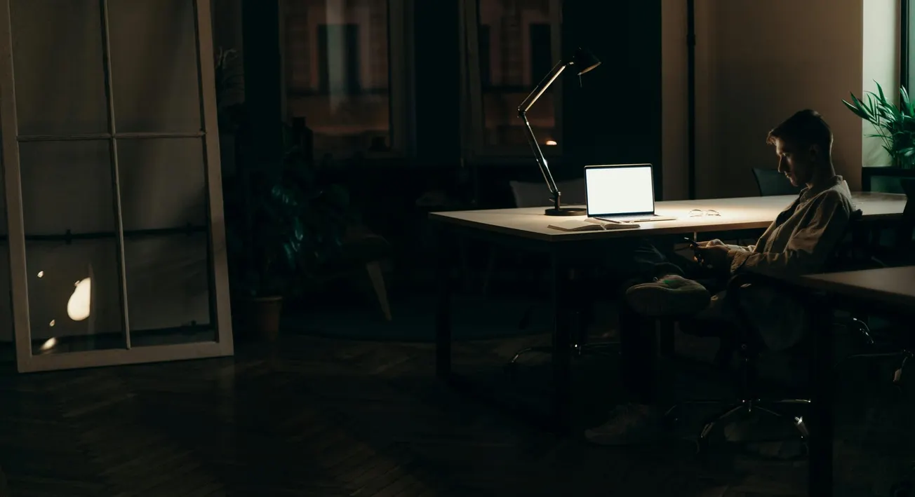 A person sits in a dimly lit room at a large desk with an open laptop, illuminated only by its screen. The atmosphere feels quiet and focused.