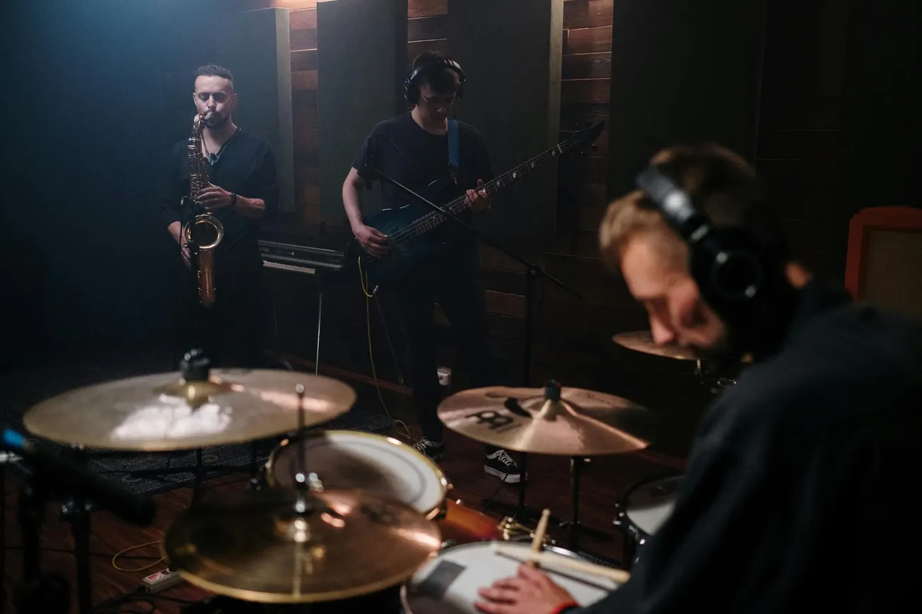 Three musicians perform in a dimly lit studio. A saxophonist and bassist play thoughtfully, while a drummer focuses intently. The atmosphere is intimate and creative.