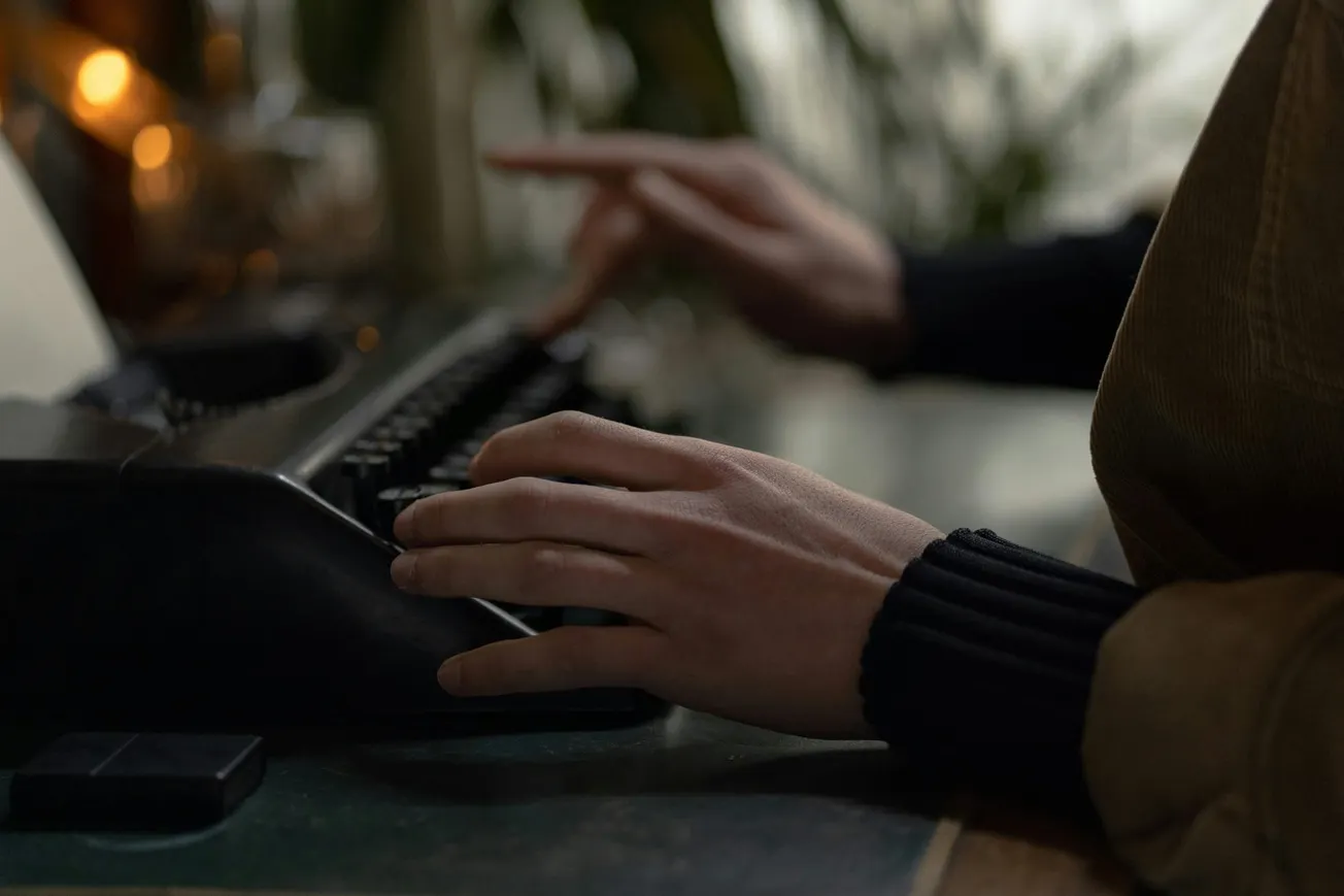 Hands typing on a vintage typewriter in dim light, creating a focused and nostalgic atmosphere. Blurred plants and warm tones in the background.