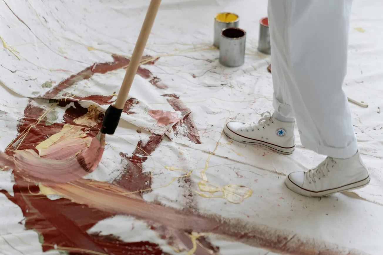 Person in white sneakers and pants painting a large canvas on the floor with abstract brown and yellow strokes, using a long brush.