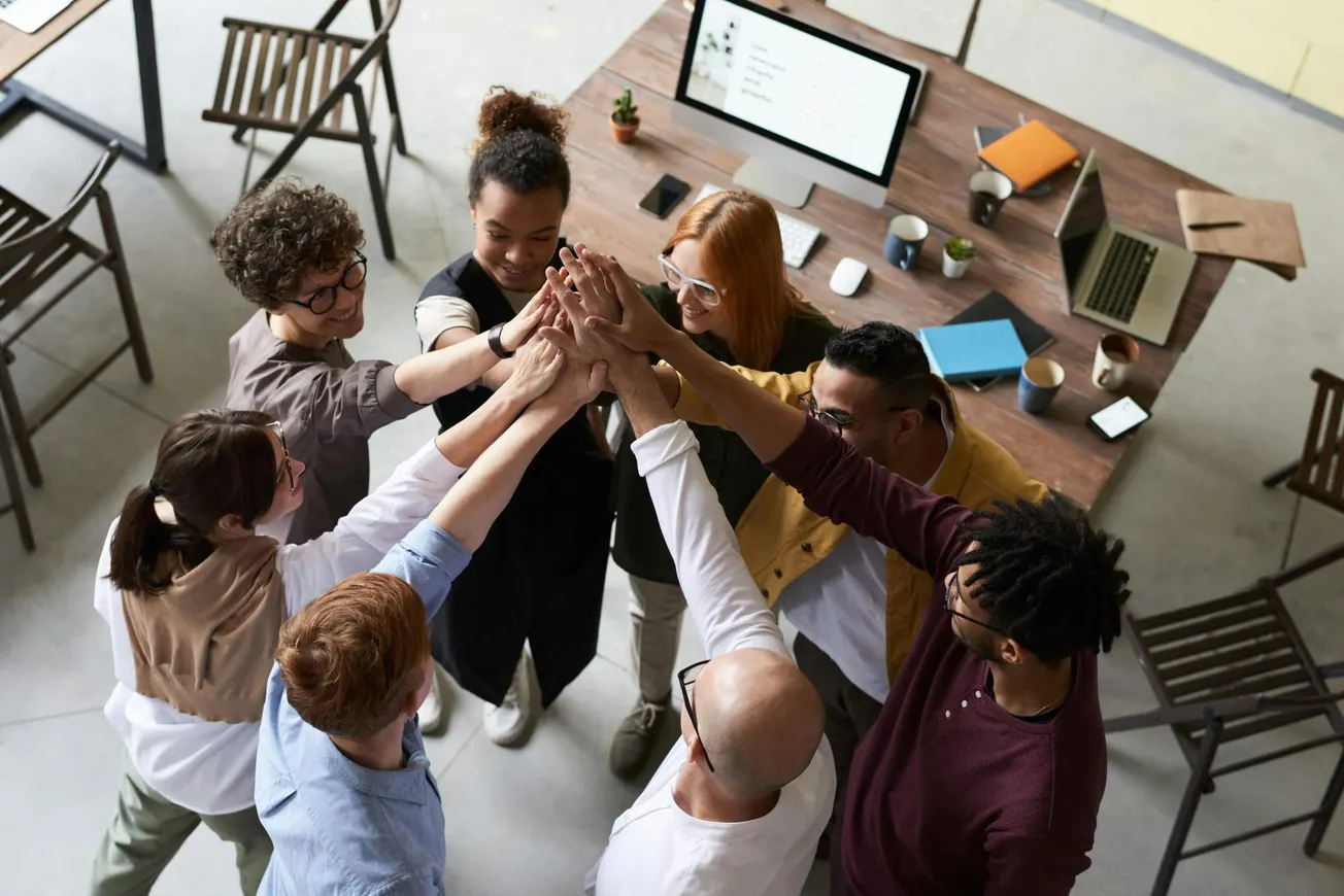 A diverse group of people stands in a circle, reaching in for a collective high-five, smiling and expressing teamwork in a collaborative office space.