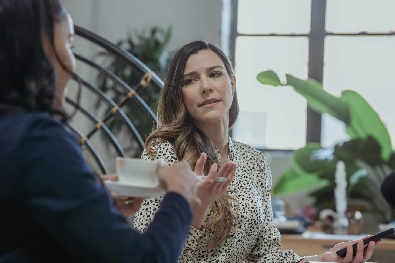 Two women are in conversation in a bright room. One holds a phone, the other a teacup. Large plants in the background.