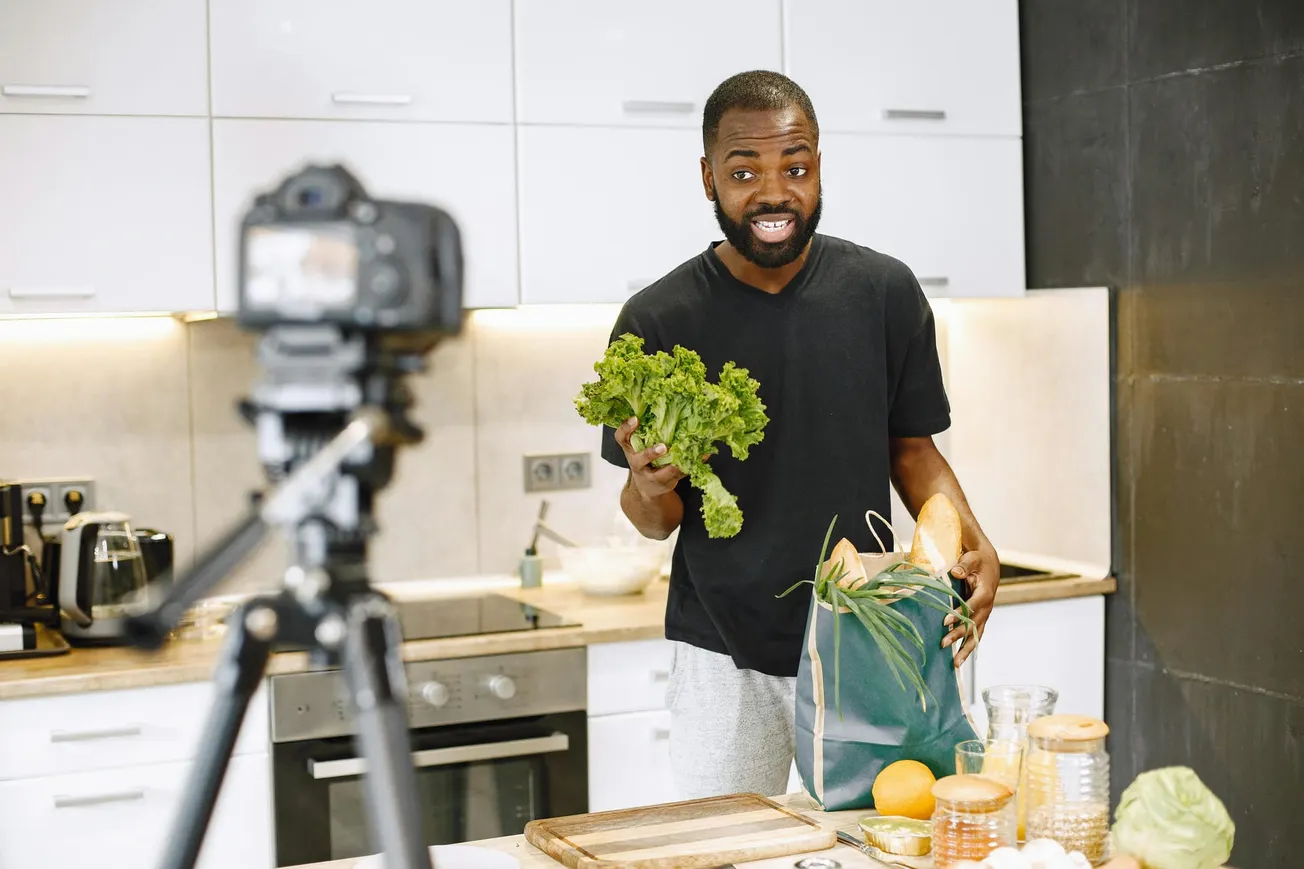 A man in a kitchen holds lettuce and a bag of groceries while smiling at a camera on a tripod, suggesting a cooking video setup.