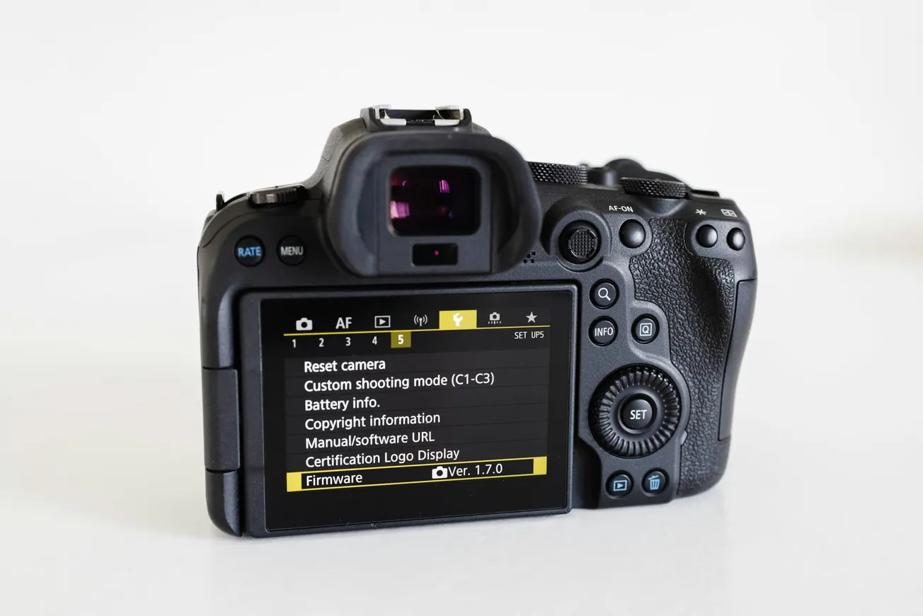 Back view of a DSLR camera displaying settings on its screen, highlighting firmware version 1.7.0. The camera rests on a white surface.