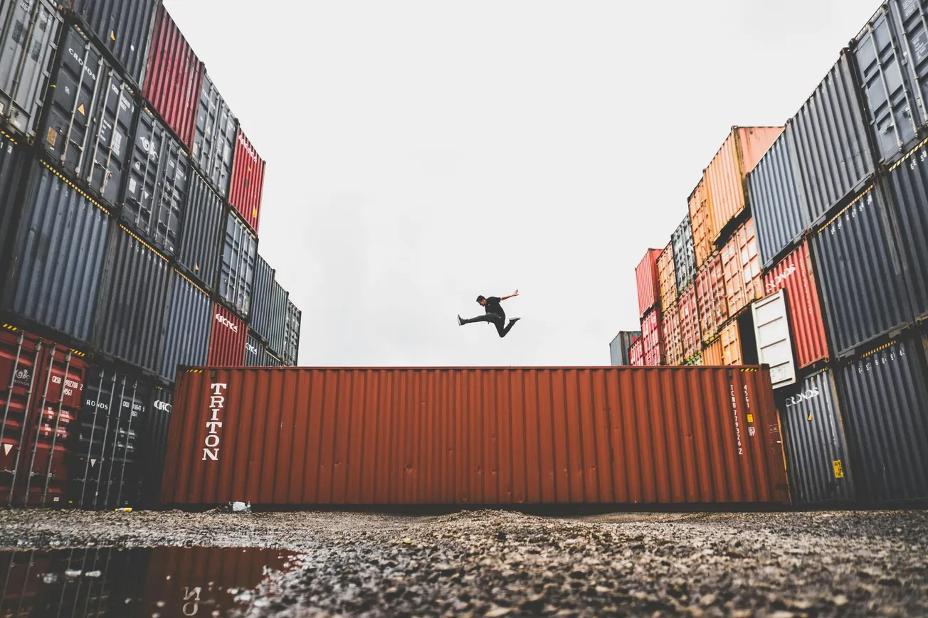 Person in mid-air, performing a dynamic jump between stacked rows of colorful shipping containers against an overcast sky, conveying energy and freedom.