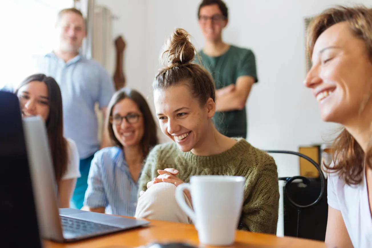 A group of five people smiles while looking at a laptop, creating a joyful atmosphere. A white mug is in the foreground on a wooden table.