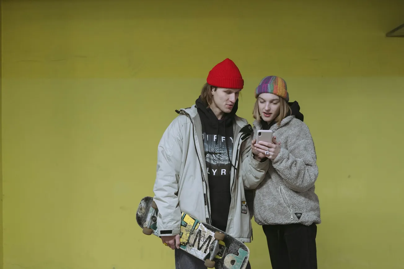 Two people in winter jackets and colorful beanies look at a smartphone intently. One holds a skateboard. Yellow background sets a casual tone.