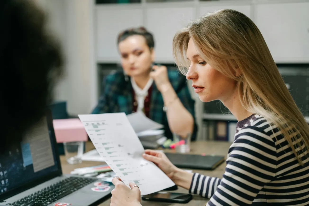 A woman in a striped shirt reviews a printed document in an office setting, with another person in the background appearing thoughtful.