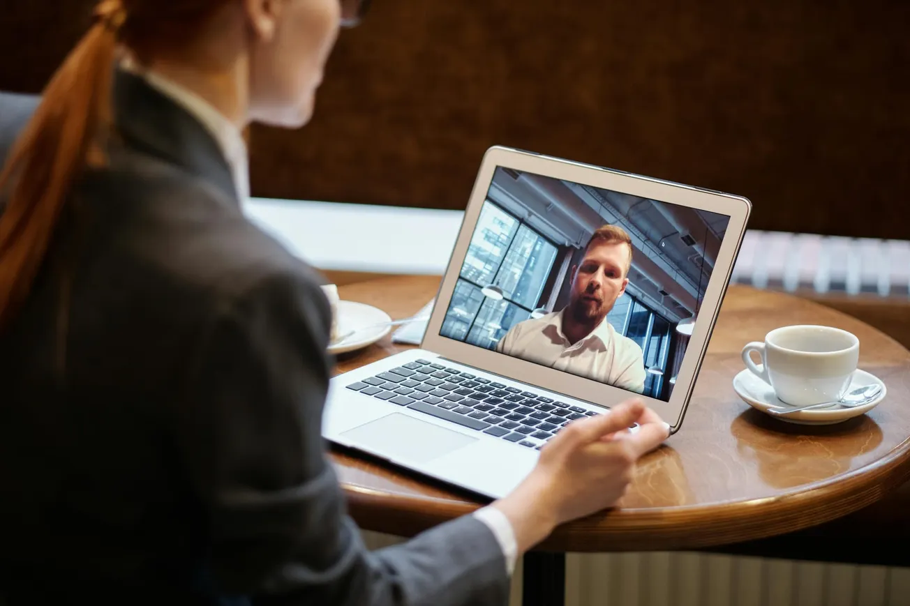 A woman in a suit watches a video call on a laptop, seated at a round table with a coffee cup. The atmosphere is professional and focused.