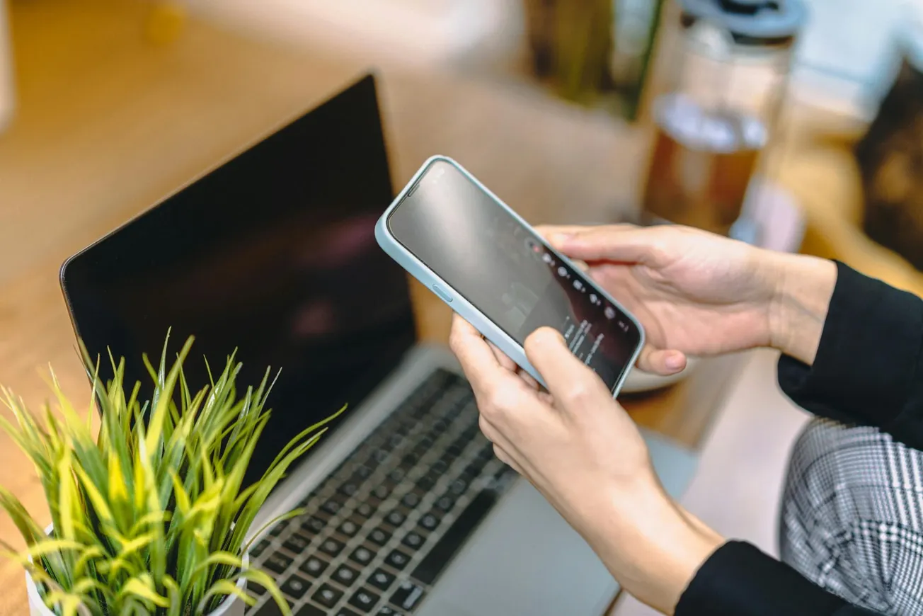 A person’s hands holding a smartphone over an open laptop on a desk. Nearby is a potted plant. The setting conveys focus and modern work.
