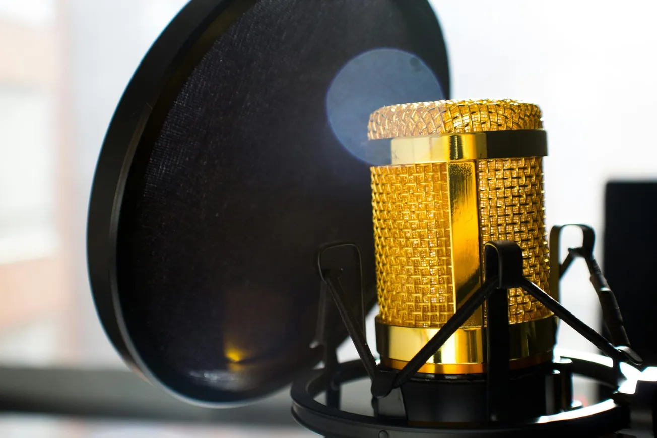 Close-up of a gold microphone in a black shock mount, next to a round pop filter. The background is softly blurred, conveying a professional studio feel.