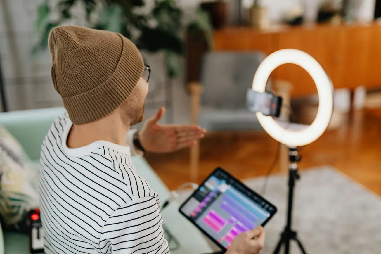 A person in a striped shirt and beanie records a video on a smartphone, using a ring light and tablet for audio control, creating a creative setup.
