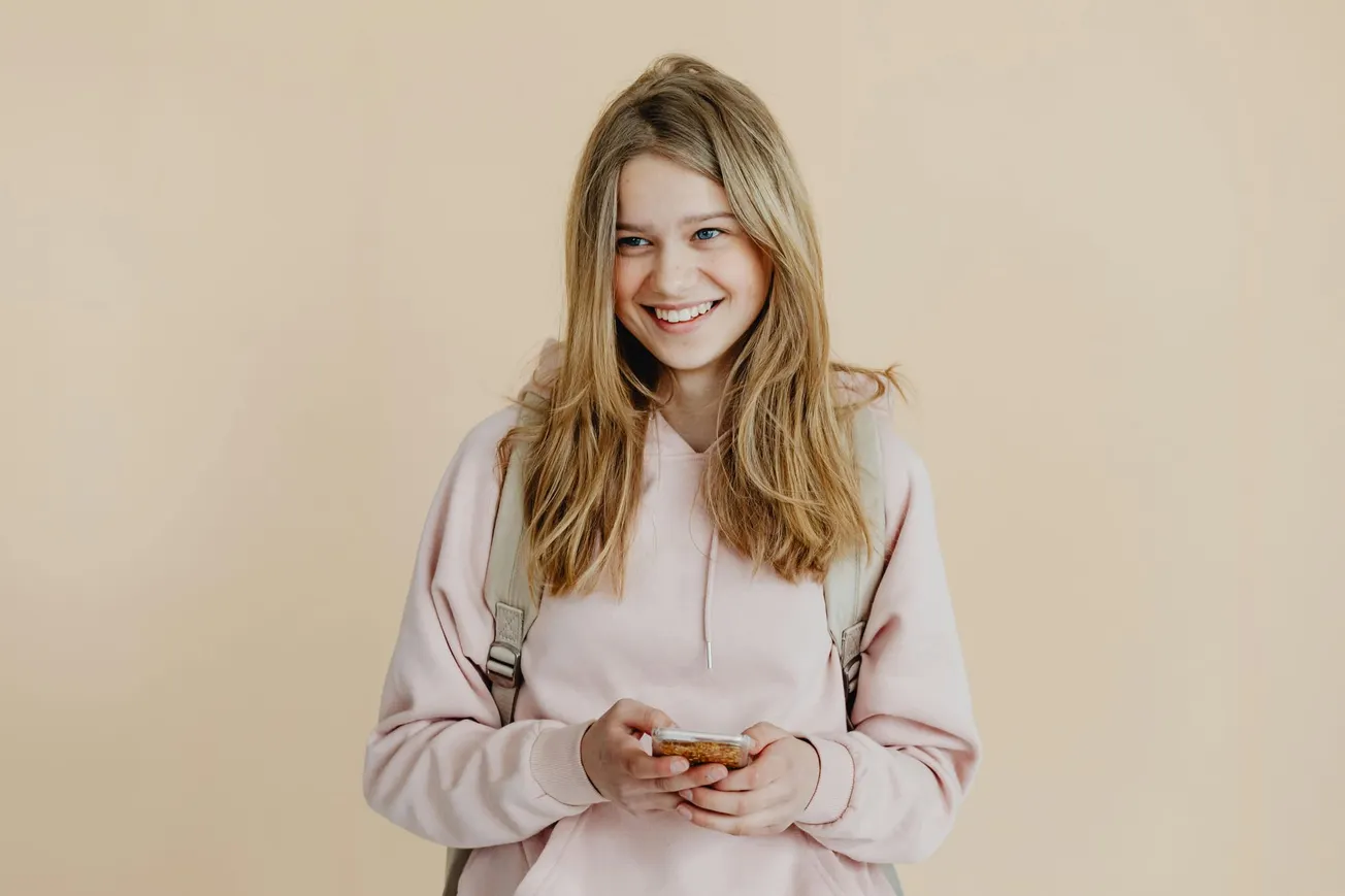 Smiling girl in a pink hoodie holds a smartphone against a beige background. She wears a backpack and exudes a cheerful, relaxed vibe.