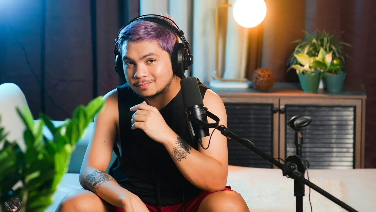 A person with purple hair and tattoos sits on a couch, wearing headphones and speaking into a microphone. The room is warmly lit with plants nearby.