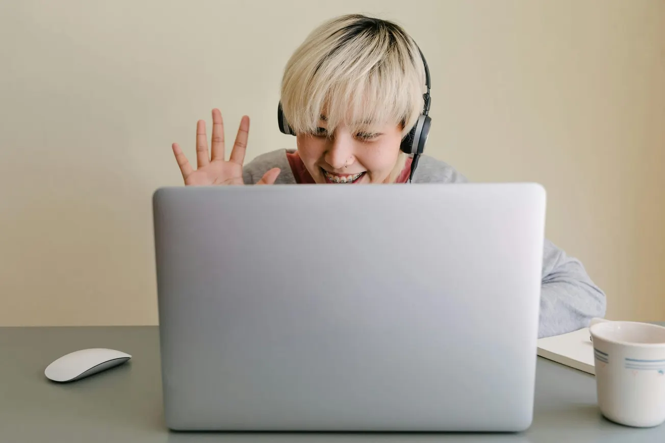 A person with short blonde hair wearing headphones is smiling and waving at a laptop screen. A coffee mug and mouse are on the desk.