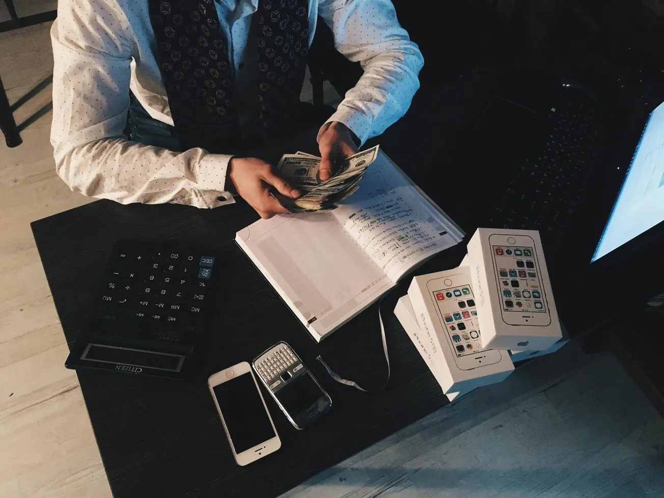 A person in a patterned shirt counts money at a desk with a notebook, stacked phone boxes, calculator, and phones. The setting conveys business activity.