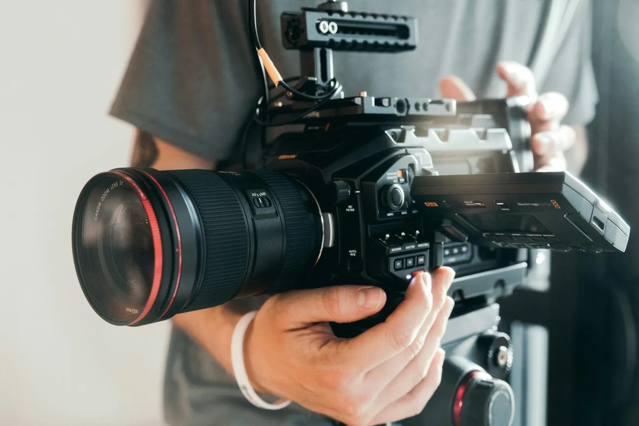 Close-up of hands holding a professional video camera with a large lens and side screen. The scene conveys a sense of focus and creativity.