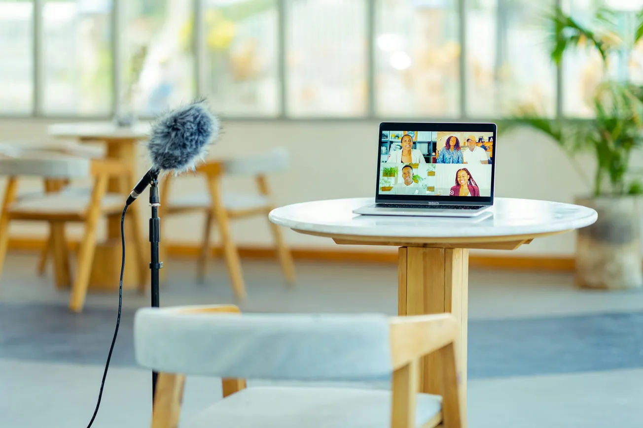 A laptop on a round table displays a video call with multiple people. A microphone on a stand is nearby. Bright, modern setting with plants.