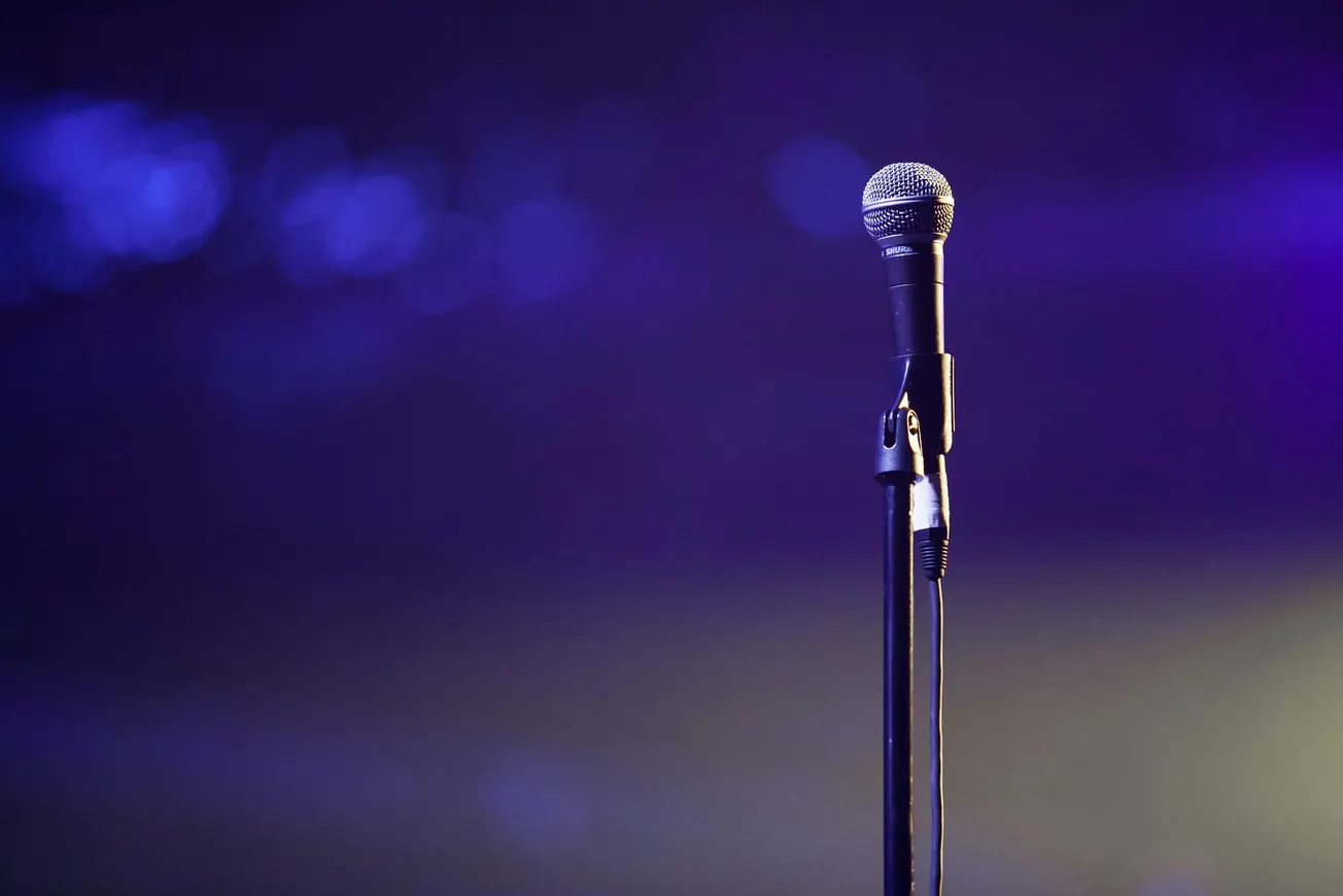 A single microphone on a stand is spotlighted against a blurred purple background, evoking a sense of anticipation and stage performance ambiance.