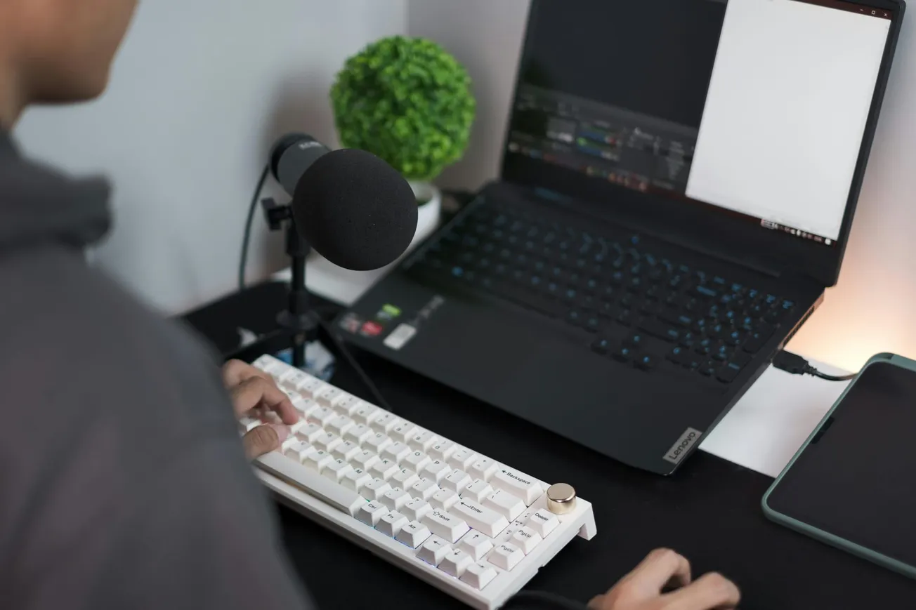 A person types on a white keyboard near a laptop with a black screen. A microphone is positioned nearby, and a small green plant adds a touch of color.