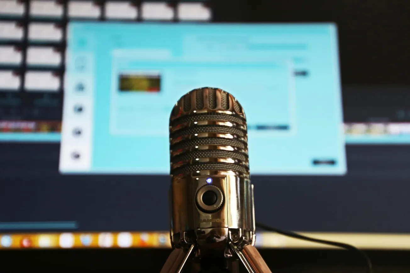 A vintage-style microphone is in focus with a blurred computer screen in the background, evoking a podcast or recording studio setting.