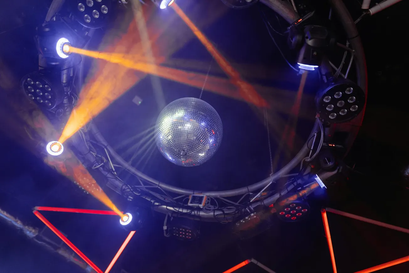 A disco ball shines amid colorful lights in a dark club. Orange and blue beams radiate from circular fixtures, creating a lively, dynamic atmosphere.