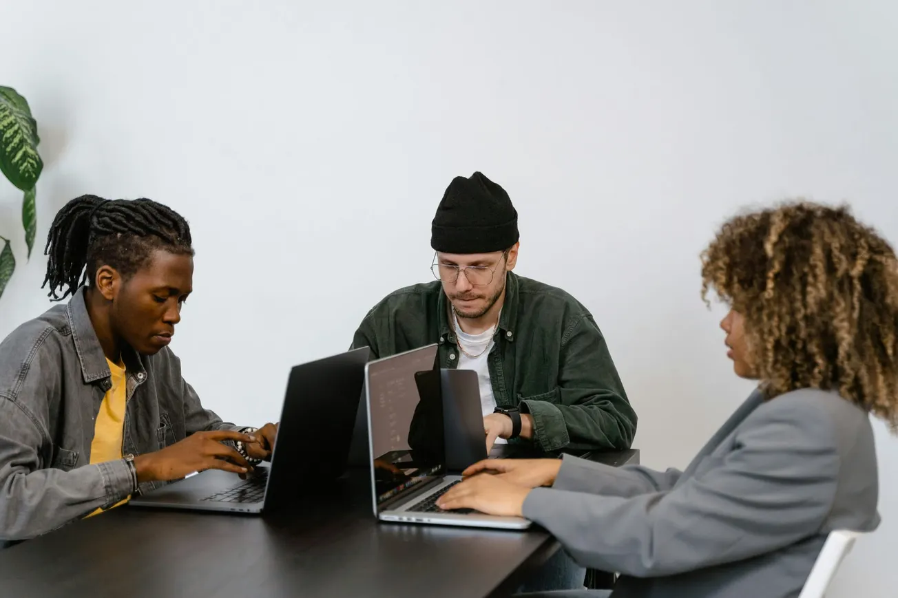 Three people sit at a table with laptops, focused on their screens. The setting is professional and collaborative, conveying a calm, studious atmosphere.