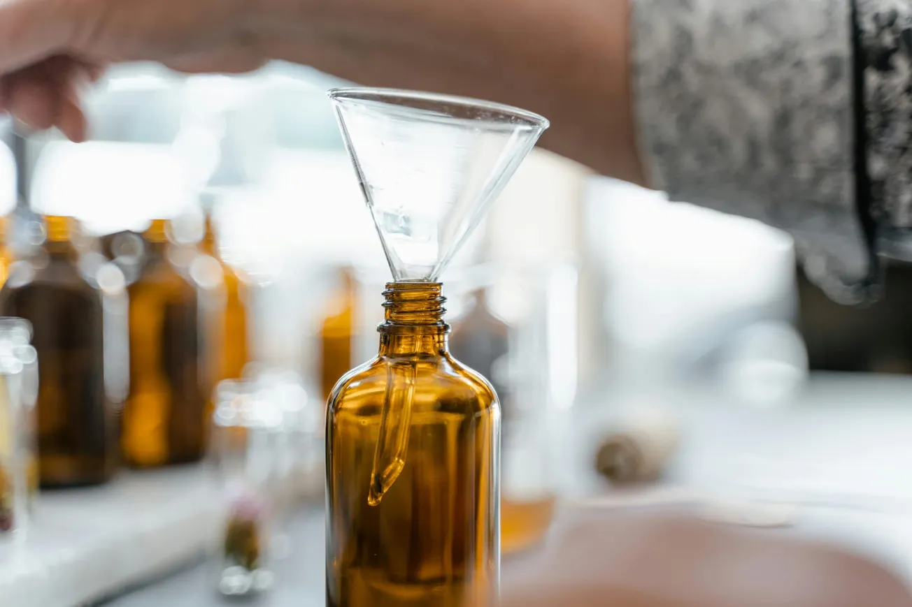A hand holds a clear funnel over a brown glass bottle, emphasizing precise, careful filling. Blurred background hints at more lab bottles, creating a focused, professional atmosphere.