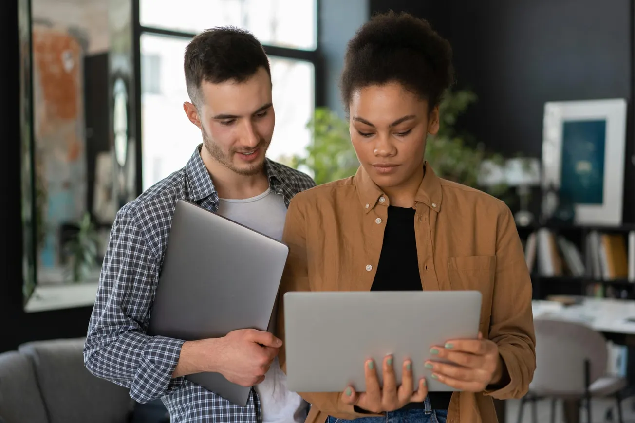 Two people in casual clothing are looking at a laptop together in a modern office. The mood is focused and collaborative.