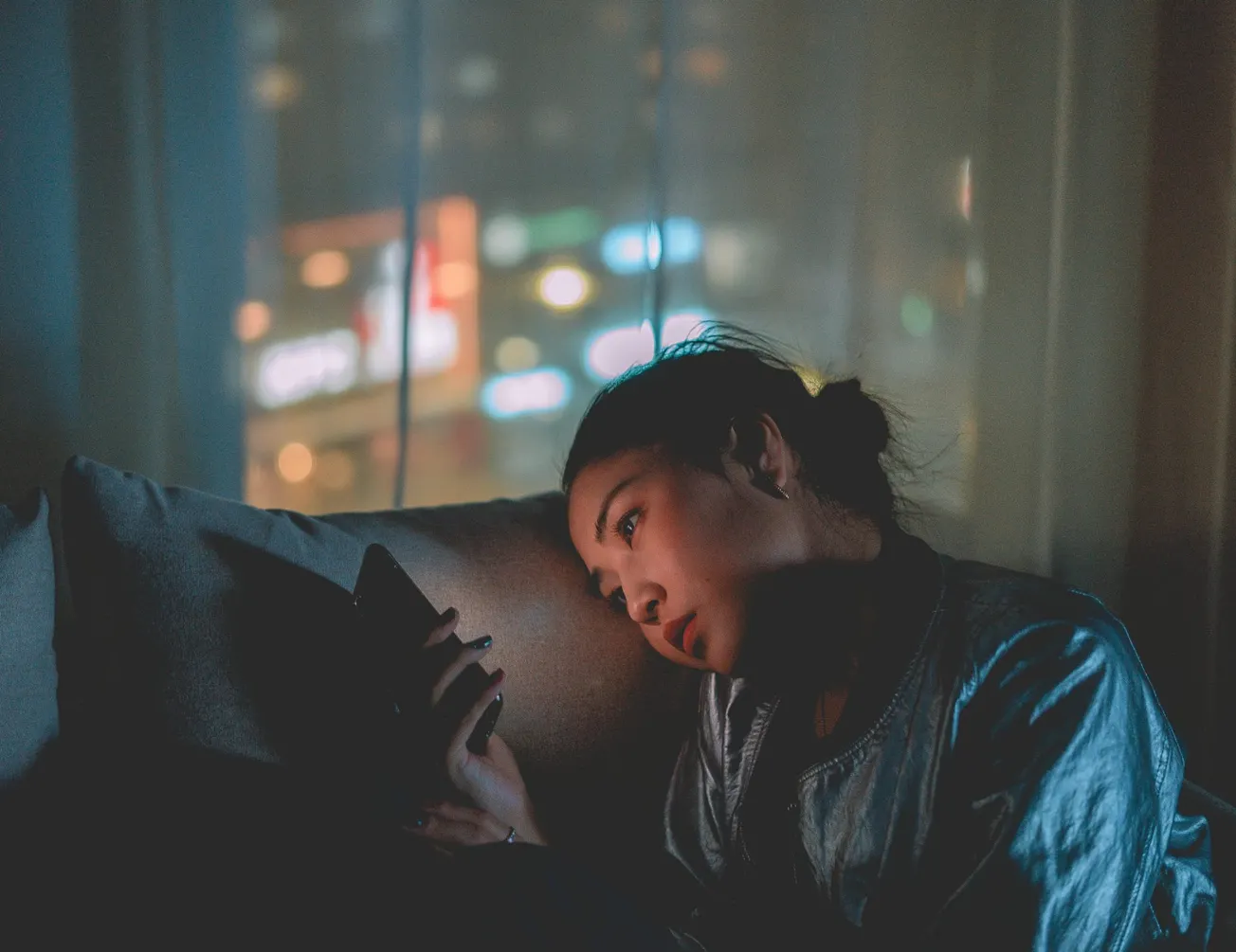 A woman leans on a pillow in a dimly lit room, absorbed in her phone. Soft city lights glow through a window, creating a calm and reflective atmosphere.