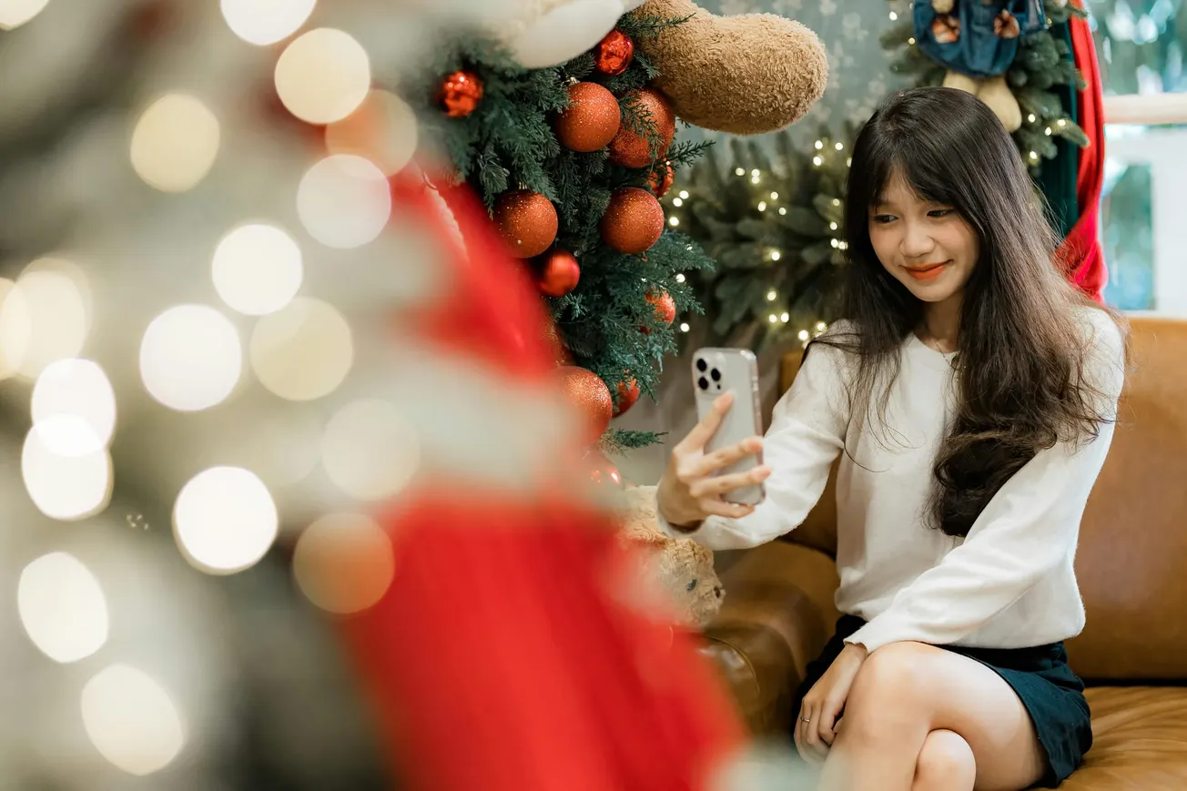 A woman sitting on a couch smiles while taking a selfie in front of a Christmas tree adorned with red ornaments. The scene is festive and joyful.