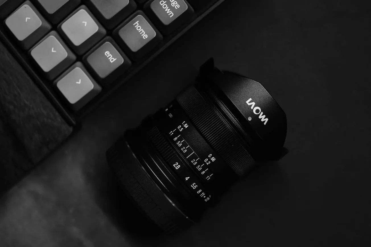 A black camera lens lies next to a keyboard on a dark surface. The keyboard keys include directional arrows and "home" and "end" labels. The scene feels minimalistic.