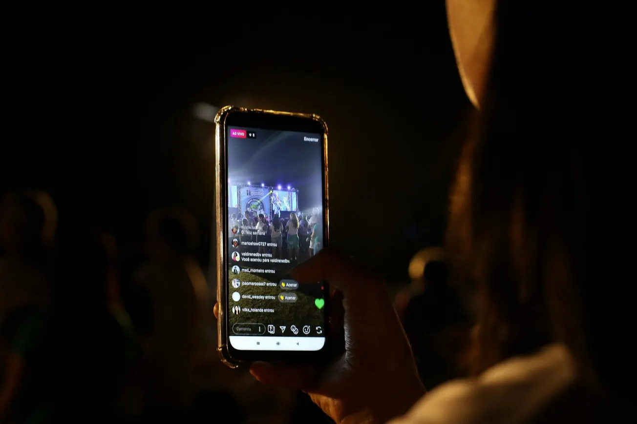 A person holds a smartphone capturing a live concert in a crowded venue. The screen displays the stage and audience with vibrant lights, conveying energy.