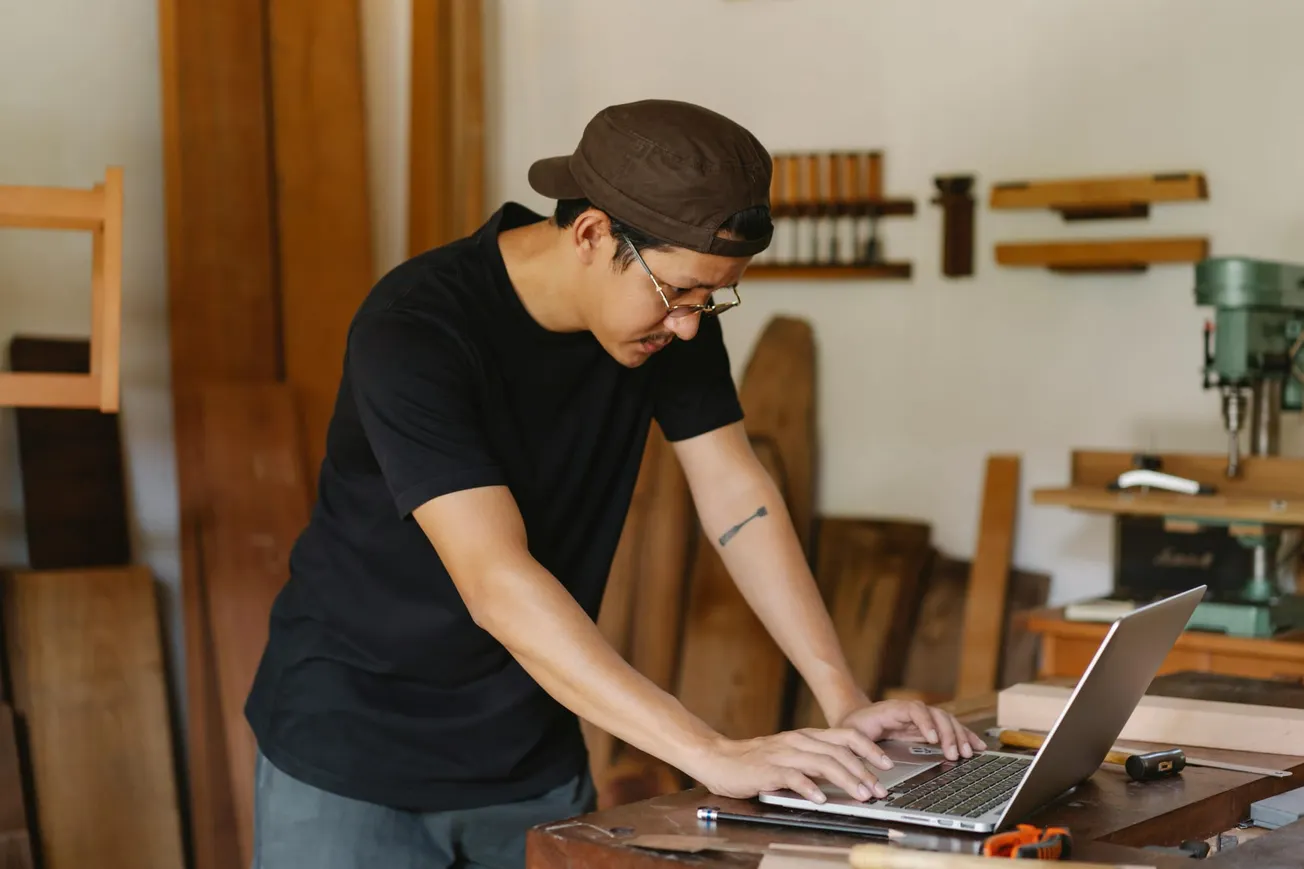 A person in a black shirt and cap focuses intently on a laptop in a woodworking shop. Tools and wooden planks fill the background, creating a busy, industrious atmosphere.