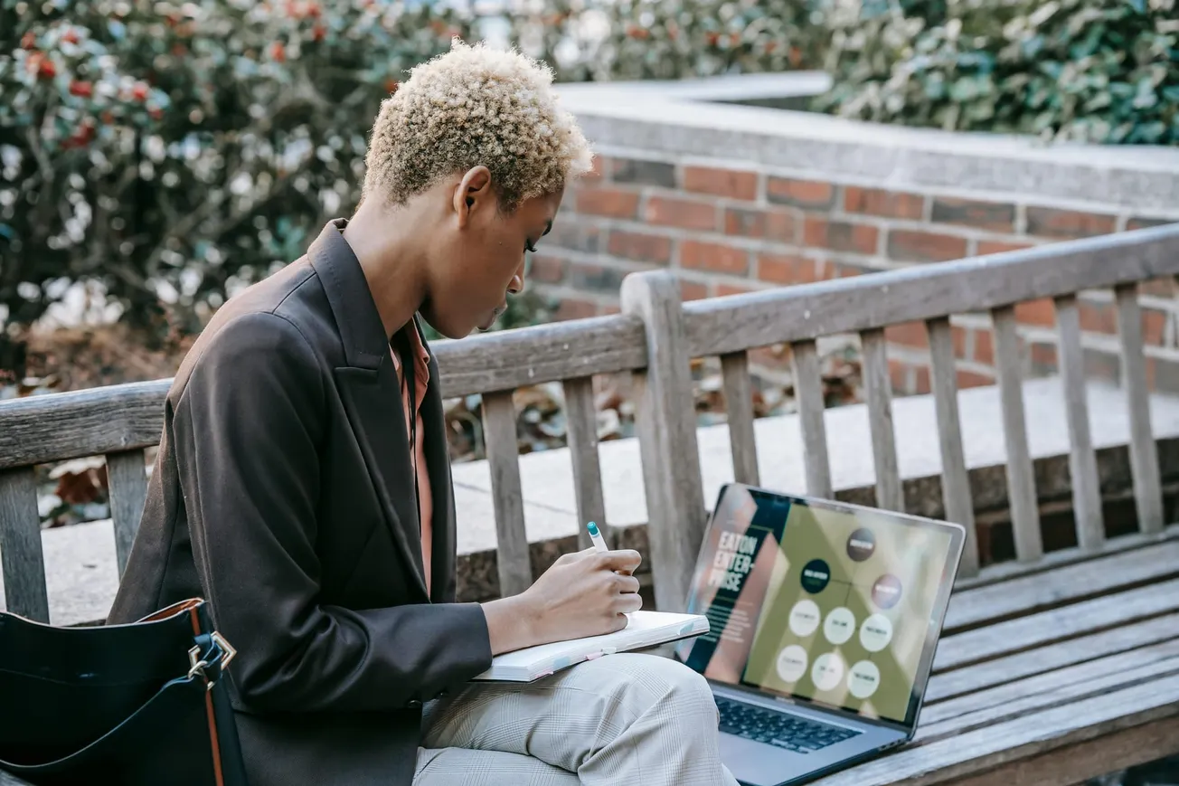 A person with short blond hair sits on a wooden bench outdoors, writing in a notebook. An open laptop beside them displays colorful charts.