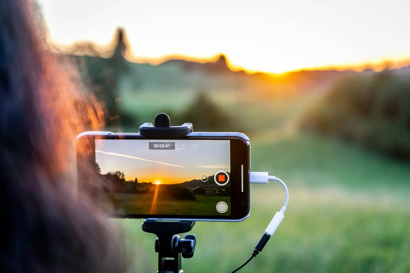 A smartphone on a tripod records a vibrant sunset over lush green fields. A cable is attached to the phone, capturing the serene, natural beauty in focus.