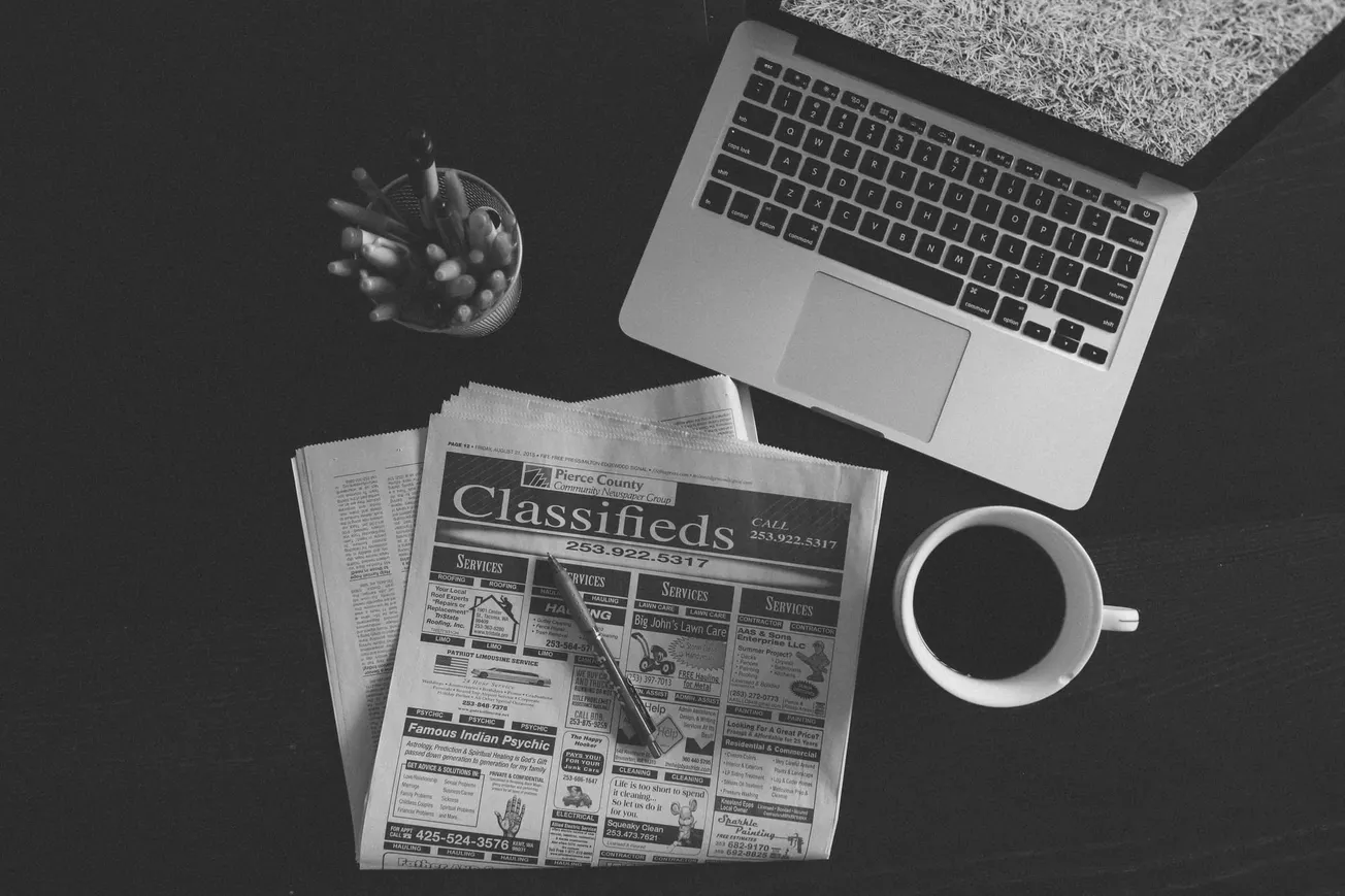 Black and white image of a laptop, a cup of coffee, and a newspaper with classifieds on a desk. A pencil lies across the newspaper, evoking a thoughtful mood.