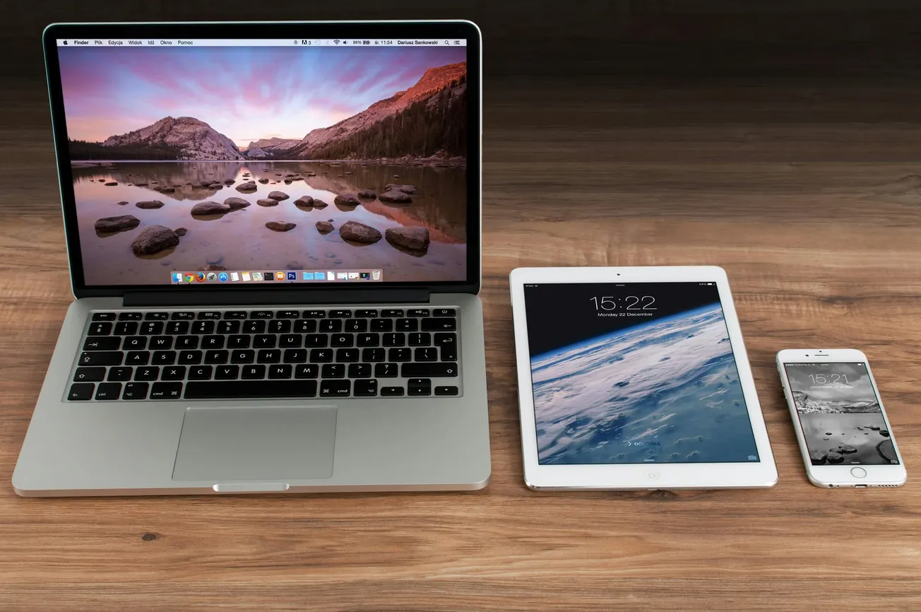Laptop, tablet, and smartphone on a wooden desk. The laptop displays a mountain landscape, and the tablet and phone show earth from space.